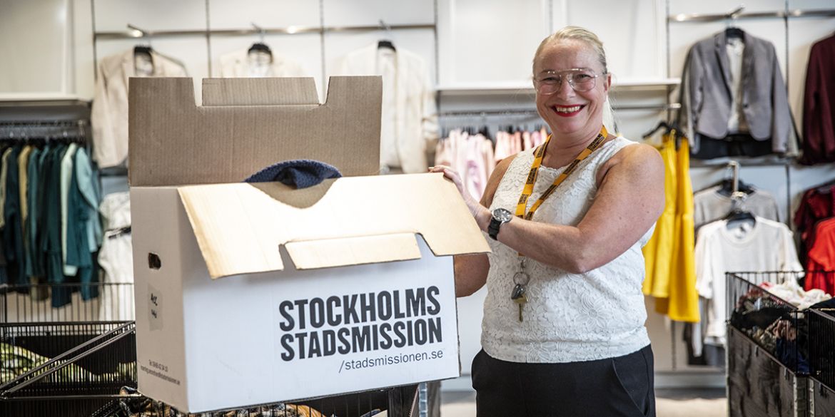 Butikschefen står med en kartong med texten Stockholms Stadsmission framför en vägg med kläder.