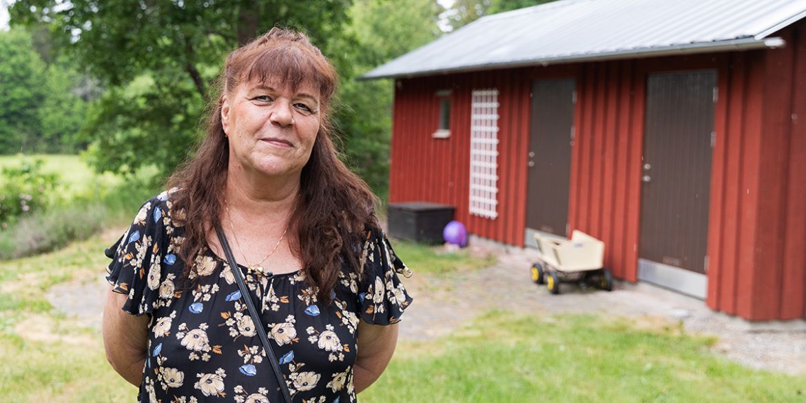 Kvinna står utomhus och tittar in i kameran med ett rött hus i bakgrunden