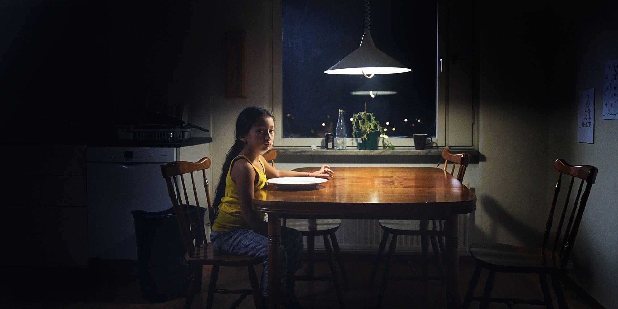 Ett liten flicka med ledset uttryck sitter ensam vid ett köksbord. Framför sig har hon en tom tallrik.