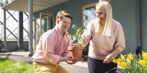 En yngre man är i en trädgård. Bredvid sitter en kvinna som håller upp en planta.