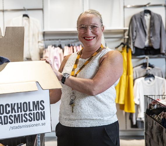 Butikschefen står med en kartong med texten Stockholms Stadsmission framför en vägg med kläder.