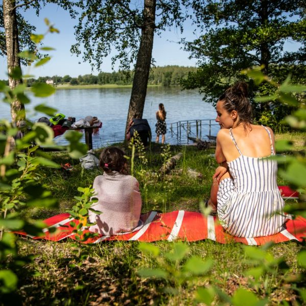 Ett barn och en kvinna sitter tillsammans på en filt med ryggen mot oss. I bakgrunden en sjö och någon som ska bada. Det är sommar. 
