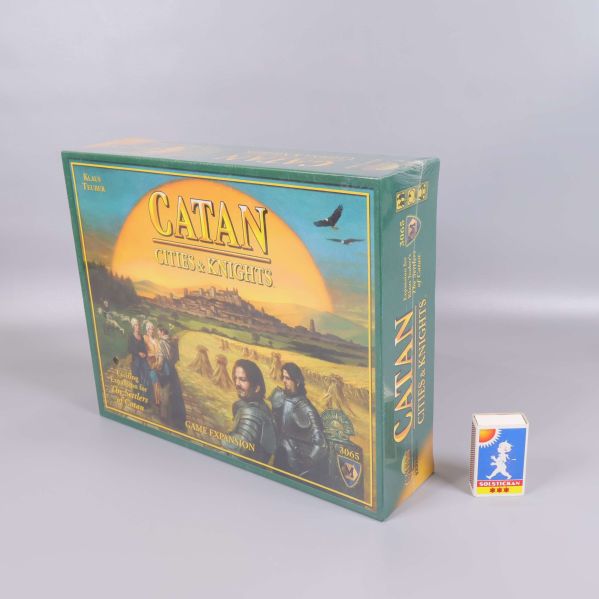 Catan Cities & Knights expansion stående bredvid tändsticksask för storleksjämförelse