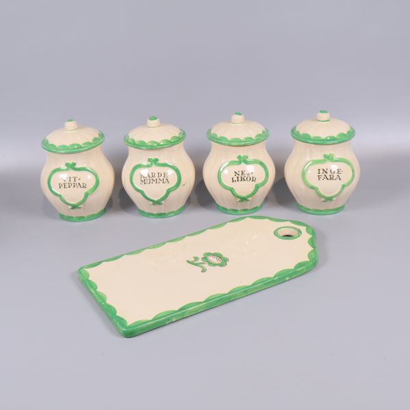 Fyra gräddvita kryddburkar med gröna detaljer och en skärbräda