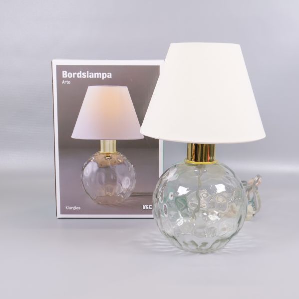 Bordslampa med klarglasfot och vit skärm framför förpackningen