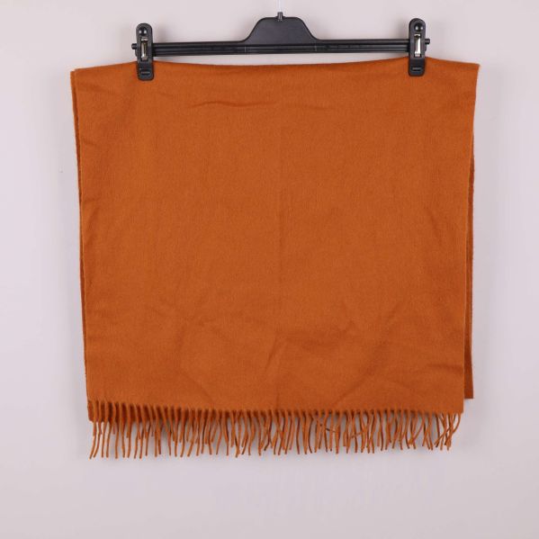 Orange halsduk med fransar upphängd mot vit bakgrund