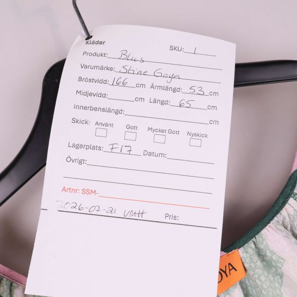 Etikett med storlek, mått och lagerplats visas mot mönstrad blus