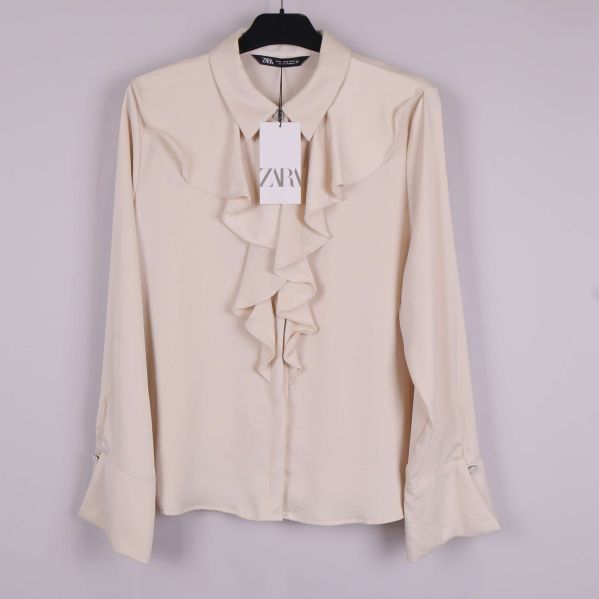 Beige blus med volang framtill och långa ärmar upphängd på galge