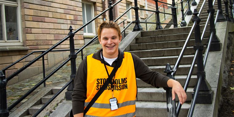 En ung man står vid en trappa med en orange Stockholms stadsmission-väst på sig. 