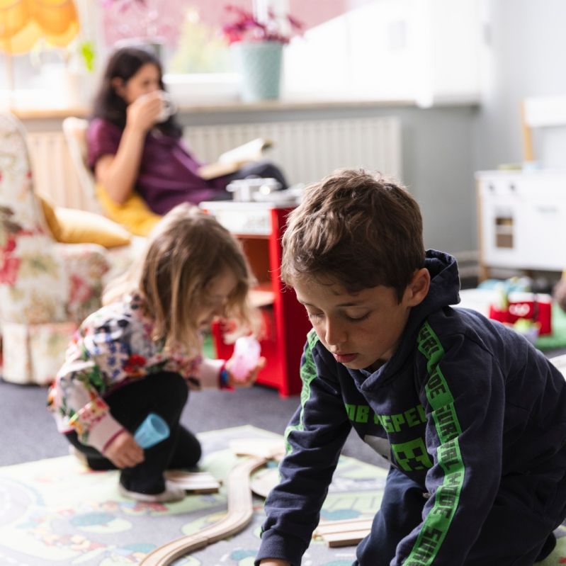Genrebild där mamma dricker kaffe i bakgrunden medan barnen ledker i förgrunden.