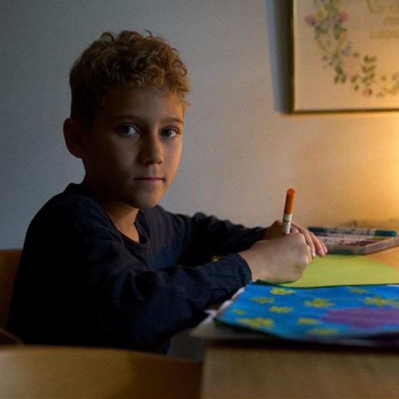 En pojke ritar en teckning och tittar in i kameran.