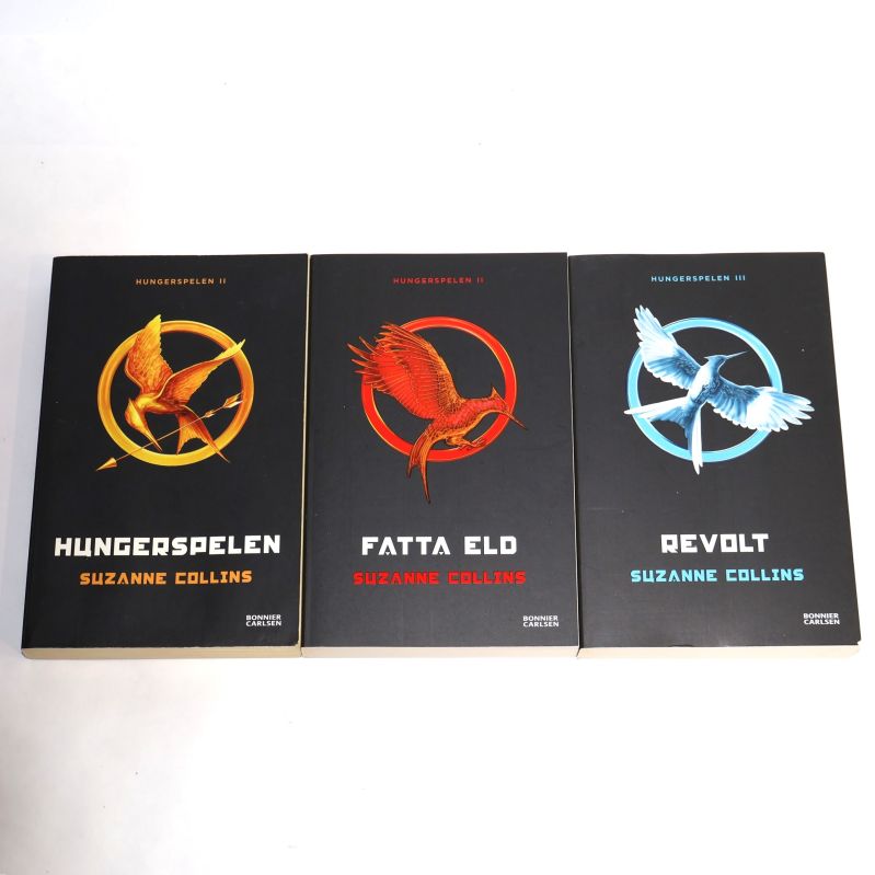 Framsidor av Hungerspelen, Fatta eld och Revolt av Suzanne Collins, alla med svarta omslag och tydliga symboler
