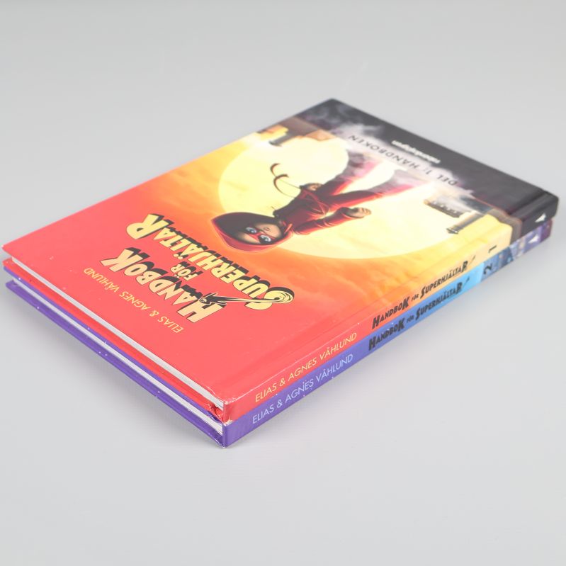Två böcker Handbok för superhjältar del 1 och 2 staplade snett mot en ljus bakgrund