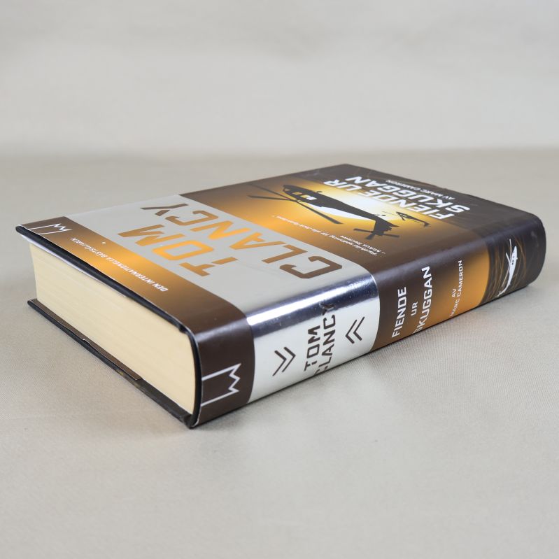 Boken Tom Clancy – Fiende ur skuggan liggande med gulddesign och hårdband