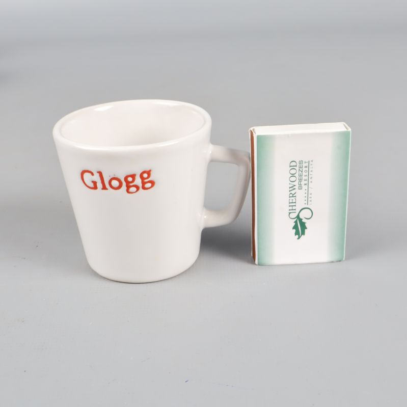 Vit glöggmugg med röd text och tändsticksask för storleksjämförelse