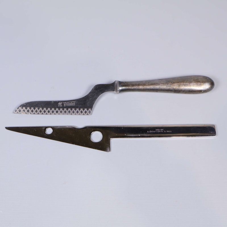 Två ostknivar med speciella bladformer liggandes bredvid varandra