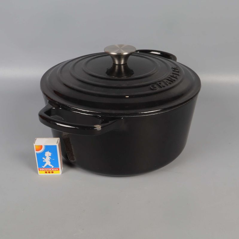 Svart gryta med lock bredvid tändsticksask för storleksreferens