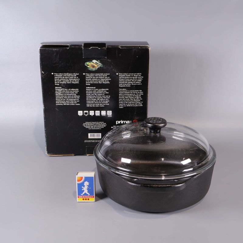Gjutjärnsgryta med glaslock framför kartongens baksida