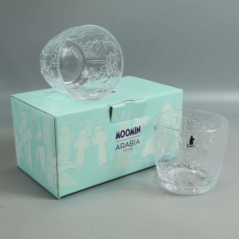 Två Mumin-glas på turkos kartong med motiv och text