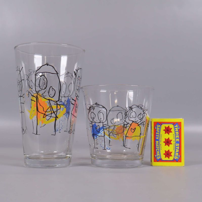 Två glas i olika storlek med färgglada figurer bredvid tändsticksask
