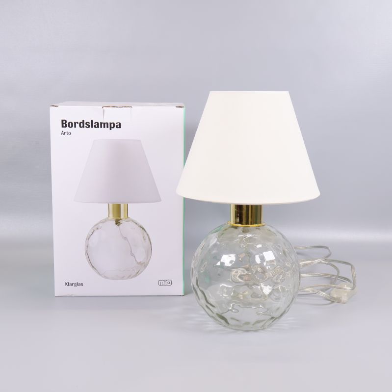 Bordslampa i glas och vit skärm framför förpackning