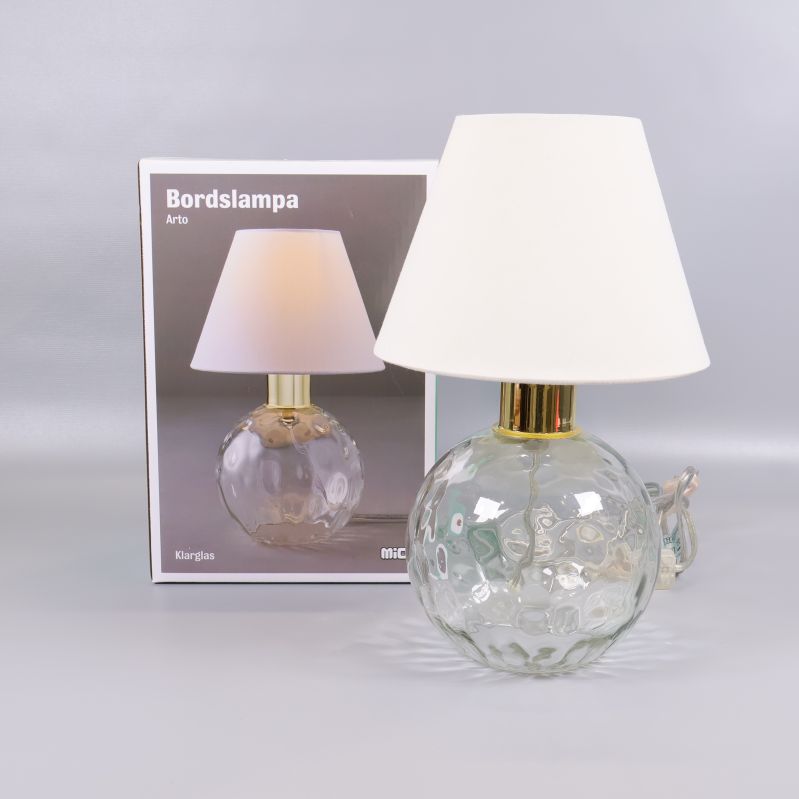 Bordslampa med klarglasfot och vit skärm framför förpackningen
