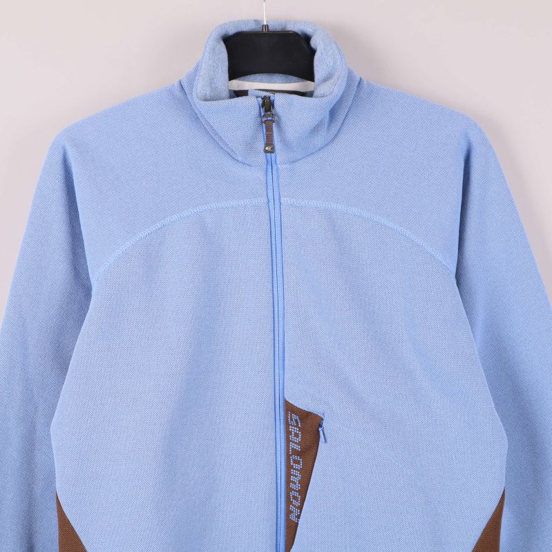 Överdel av ljusblå jacka med dragkedja och Salomon-märke vid fickan