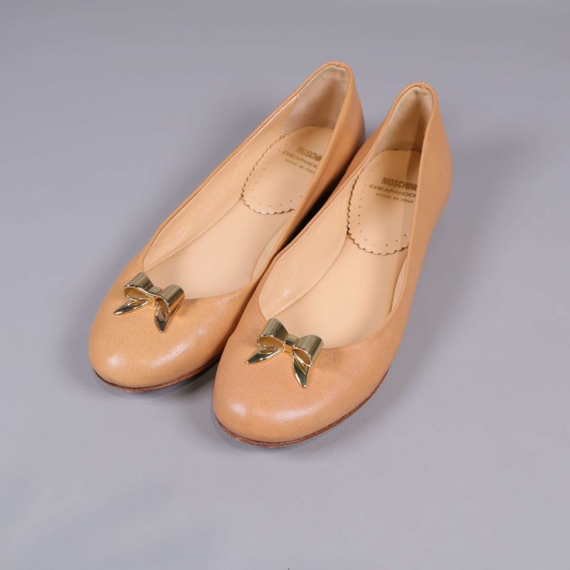 Beige ballerinaskor med guldfärgad rosett på grå bakgrund