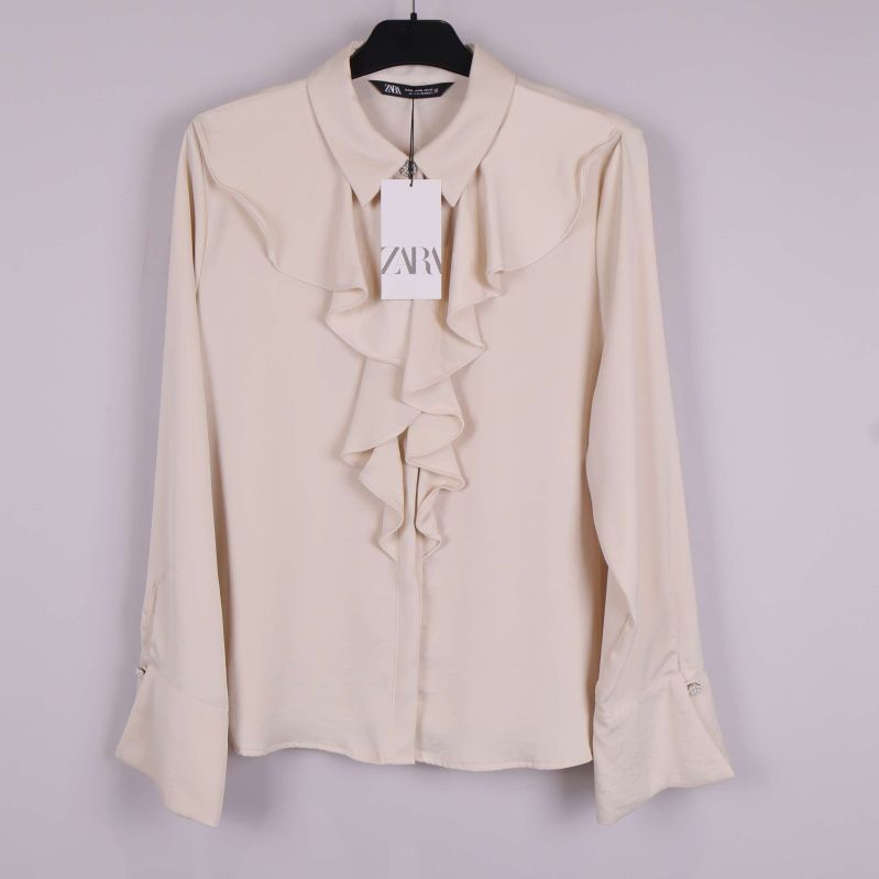Beige blus med volang framtill och långa ärmar upphängd på galge